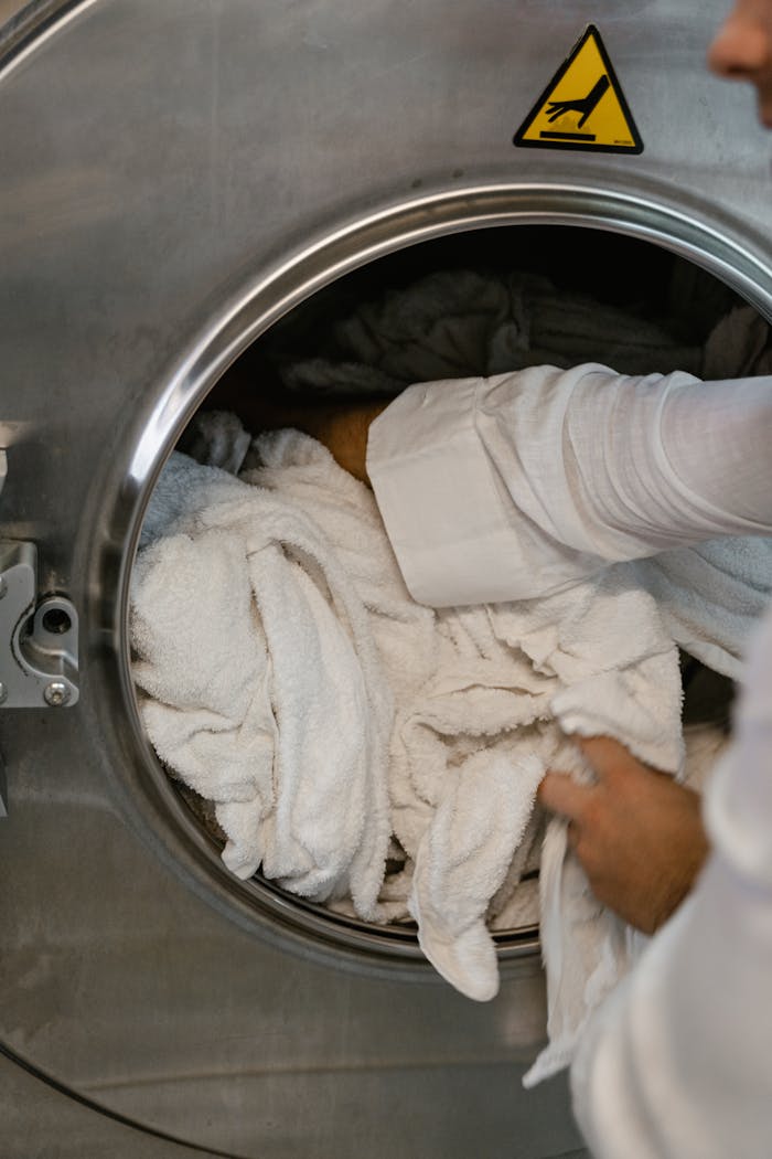 core-values Close-up of hands loading white towels into a large industrial washing machine at a laundromat.